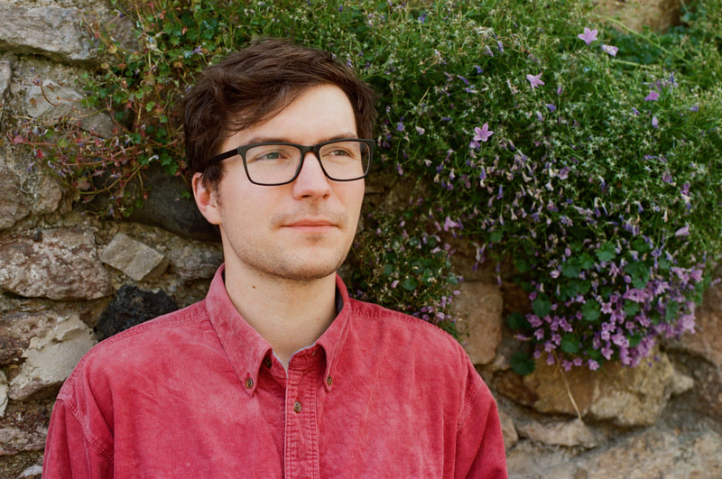 Image of Iestyn Tyne. Tyne is a white man with dark hair and glasses. He is wearing a rose red coloured shirt and is pictured with head and shoulders visible. There is a wall behind him and a climbing plant full of green foliage and pale purple flowers. 