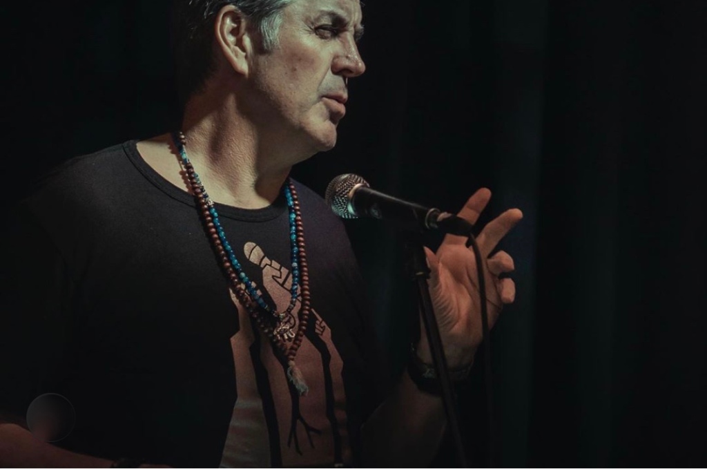Simon Maddrell stands at a mic wearing beads and a dark T-shirt with a graphic design.