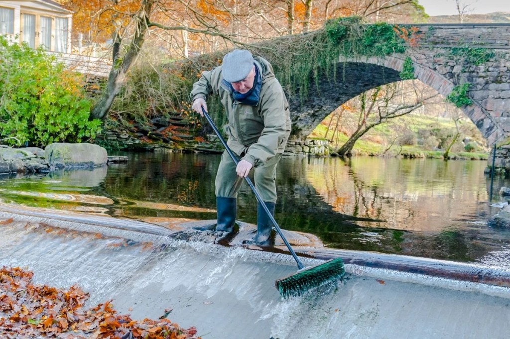 A white man in overalls and a cap stands in the water of the rover with a sweeping brush, presumably brushing leaves out of the course of the river