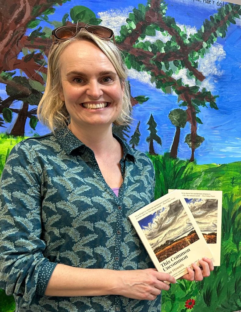 Rae Howells poses with copies of the poetry collection This Common Uncommon with a bright mural of trees and vegetation behind.