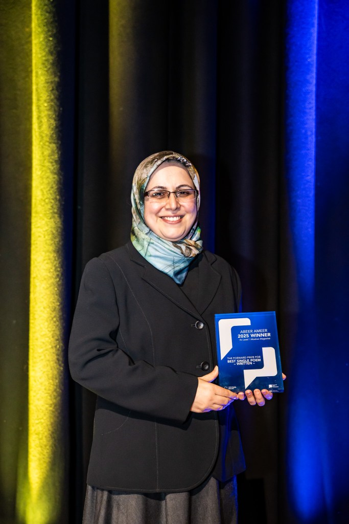 Abeer Ameer wears headscarf and black formal jacket with glasses holding the Forward Prize - a blue and white glass award. 