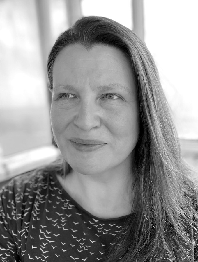 A close-up portrait of a white woman with long hair, wearing a patterned top. She has a thoughtful expression and is looking slightly off-camera.