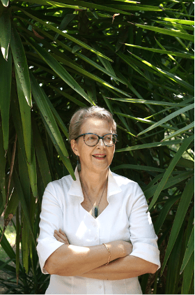 Anne Eyries is a white woman with glasses standing confidently with her arms crossed, wearing a white blouse, in front of lush green leaves.