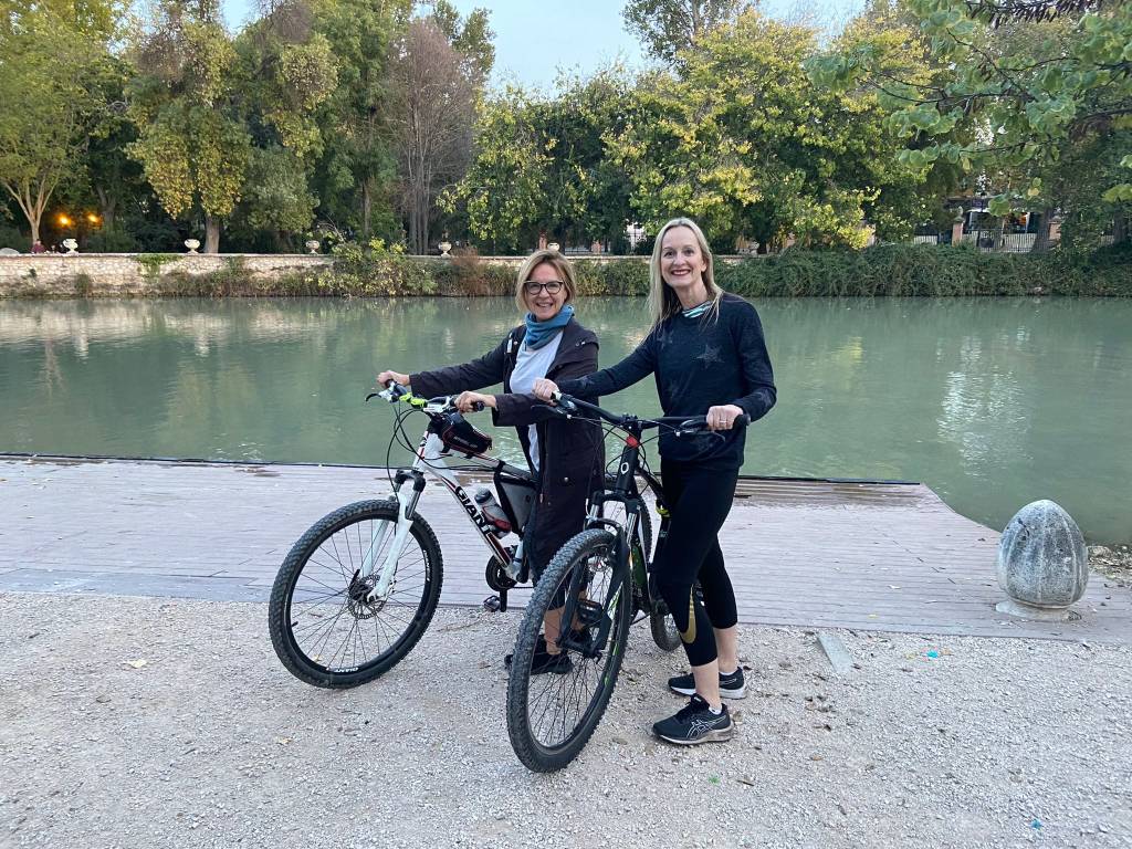 Two white women - Lisa and Ben - standing with their bicycles on a path by a river, surrounded by trees.