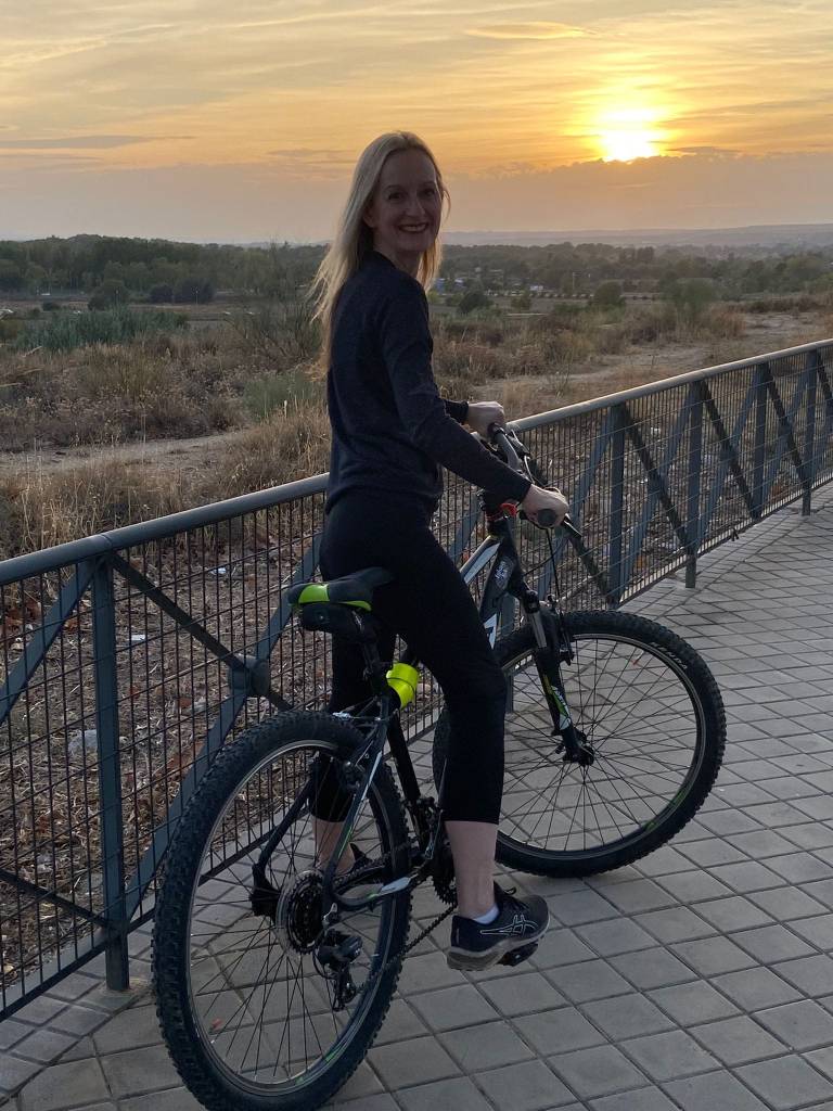 Lisa Kelly is a white woman smiling while sitting on a bicycle at sunset, with a scenic landscape in the background.