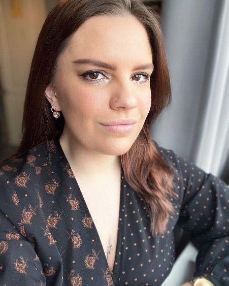 A woman with long brown hair wearing a black patterned blouse, sitting by a window with natural light