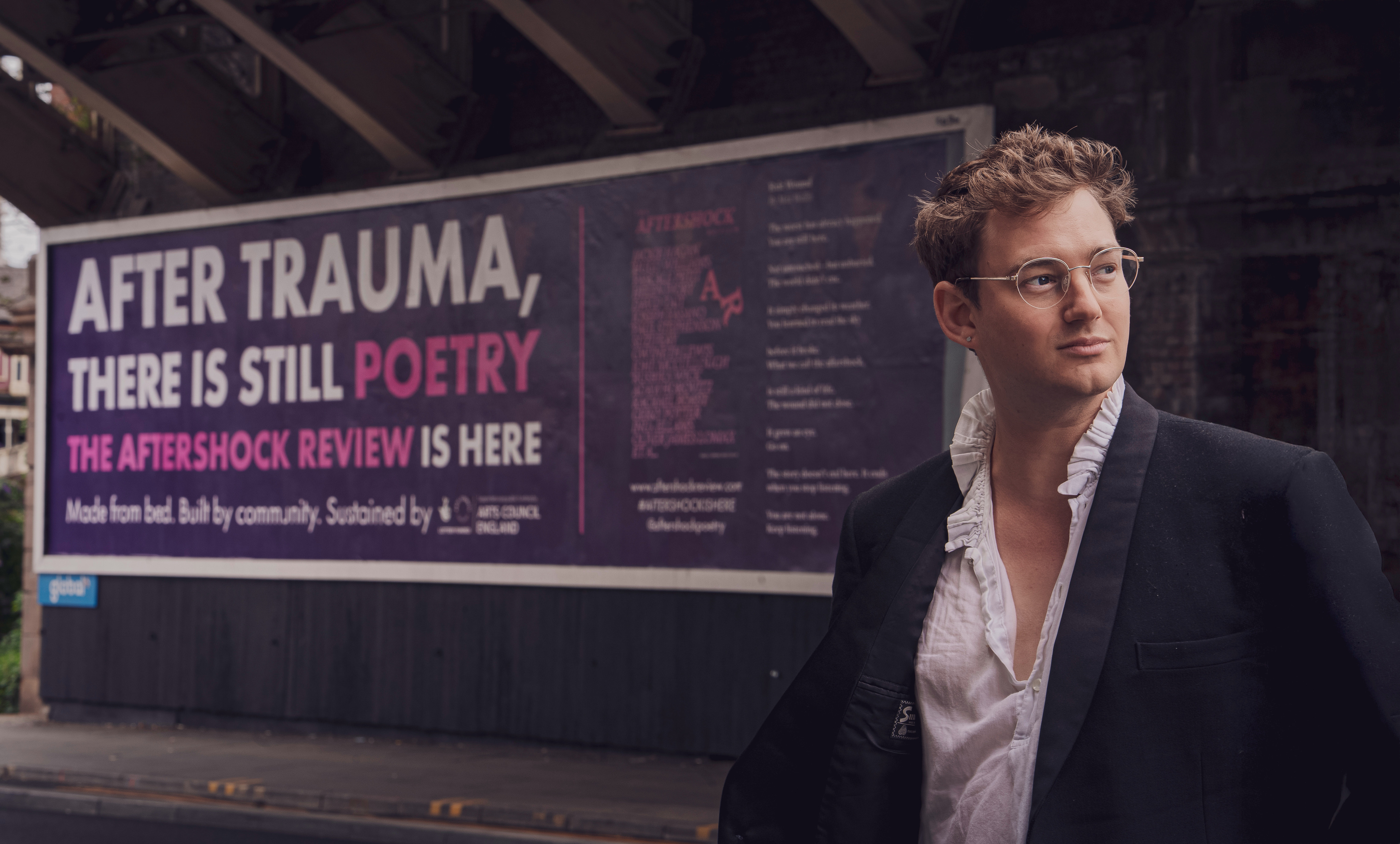A person with glasses and ruffled shirt stands near a billboard that reads 'After trauma, there is still poetry. The Aftershock Review is here'