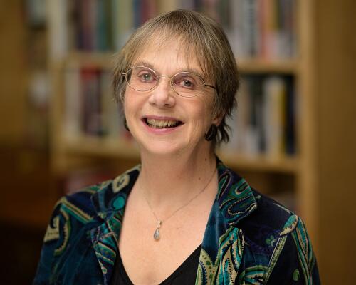 A smiling person with short hair and glasses wearing a patterned green and blue jacket in front of blurred bookshelves