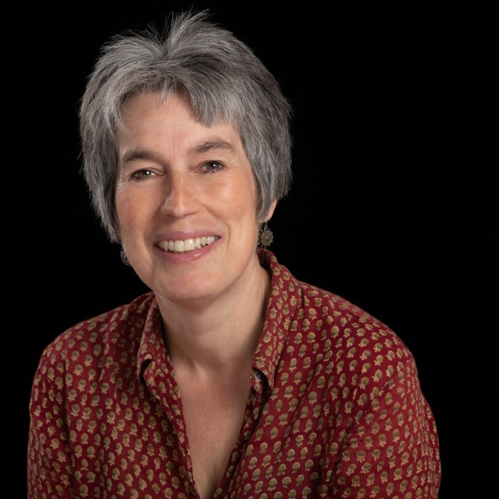 Portrait of a smiling woman with short grey hair against a black background. She wears a red patterned shirt and small earrings, and faces the camera with a relaxed, friendly expression.
