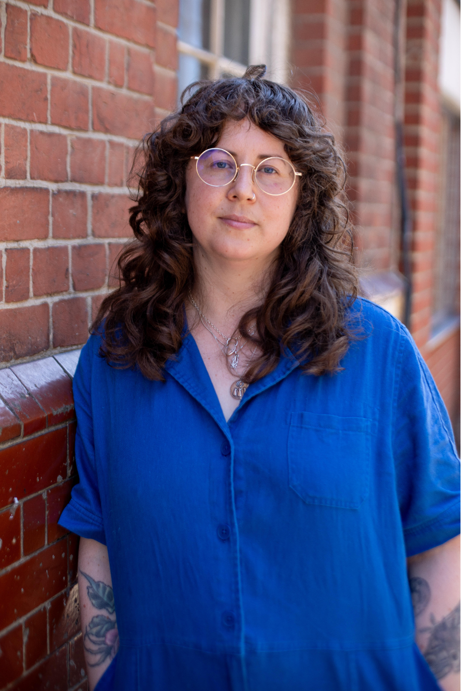 A person with curly hair and glasses stands casually against a brick wall, wearing a blue shirt and showing visible tattoos on their arms.