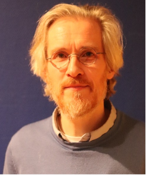 A man with blonde hair and beard wearing round glasses, a blue sweater, and a white shirt, standing against a dark blue background