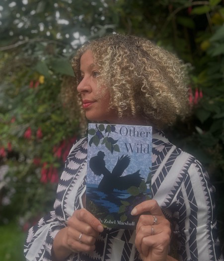 image Person holding the book 'Other Wild' in a garden with greenery and red flowers