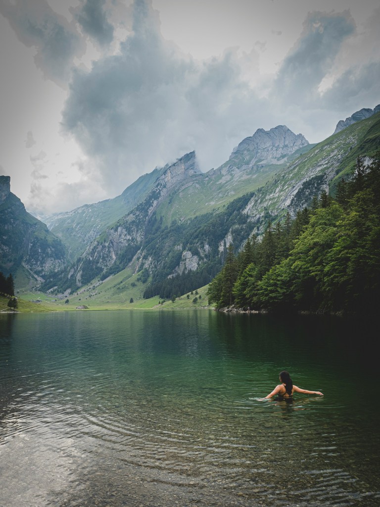 A woman stands in a green lake surrounded by rugged mountains