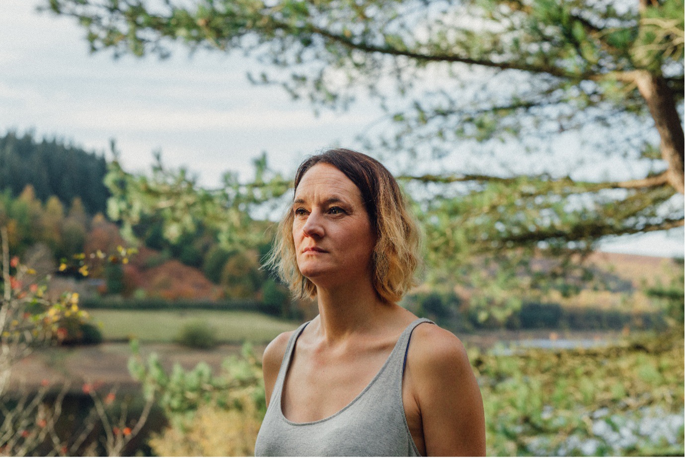 Genevieve Carter A white woman with shoulder-length hair looking thoughtfully into the distance outdoors with trees and hills in the background