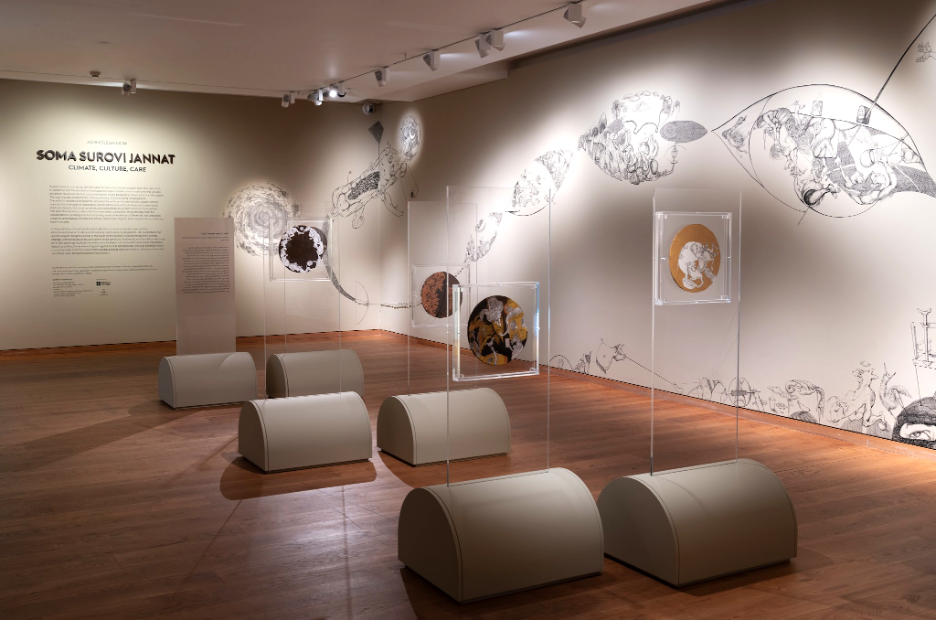 Art gallery exhibit titled Soma Surovi Jannat featuring cylindrical pedestals with framed circular artworks and a large mural with intricate black and white drawings on the wall