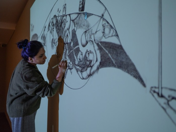 An artist drawing detailed black ink figures on a white wall in a dimly lit room